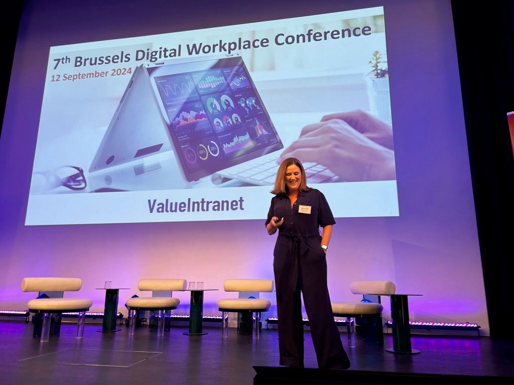 A woman (me) is on stage, holding a slide clicker. She has brown hair and is wearing a dark blue jumpsuit. She is smiling.