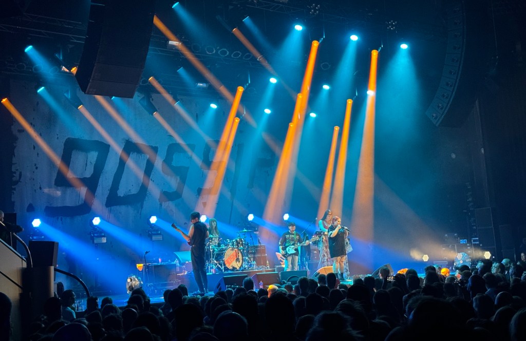 The band Gossip on stage at the Tivoli Vredenberg in Utrecht. Beth Ditto is wearing a black sprakly dress. The word GOSSIP is across the back of the stage in graffiti-like writing. There are blue and yellow lights on the stage.
