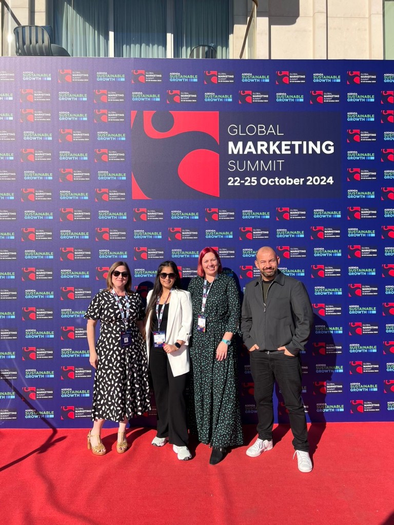Four people stand in front of a branded sign for the Global Marketing Summit
