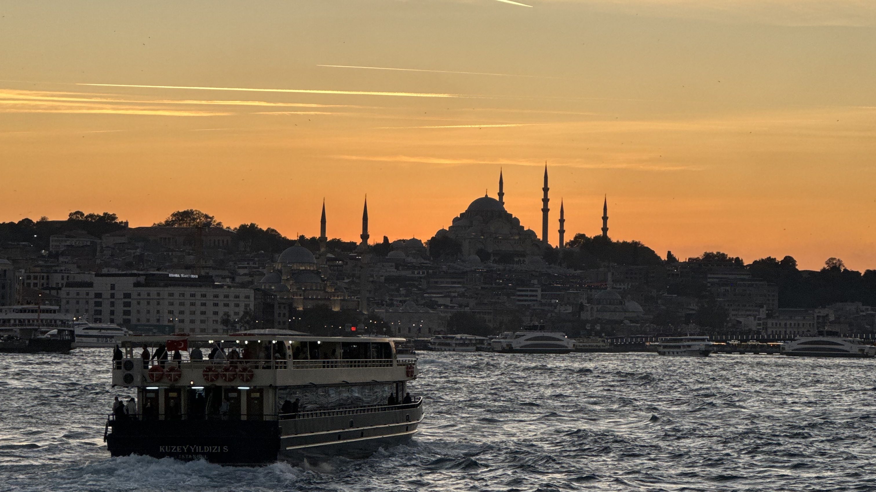A. view of the Sulemaniye Mosque in Istanbul at sunset. The river is in the foreground and there's a ferry sailing on it.
