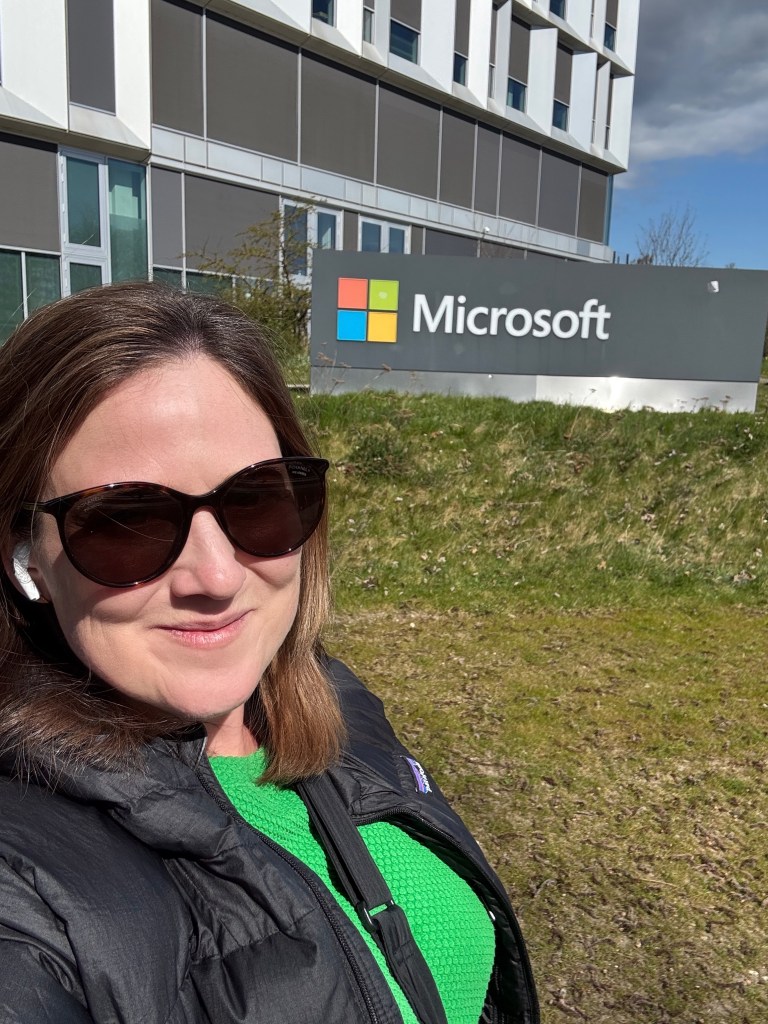 A white woman with brown hair stands in front of a Microsoft sign. She is wearing sunglasses, a bright green top and a black jacket,