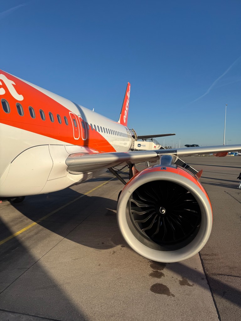 The engine and side of an orange and white EasyJet plane, on the tarmac with a clear blue sky