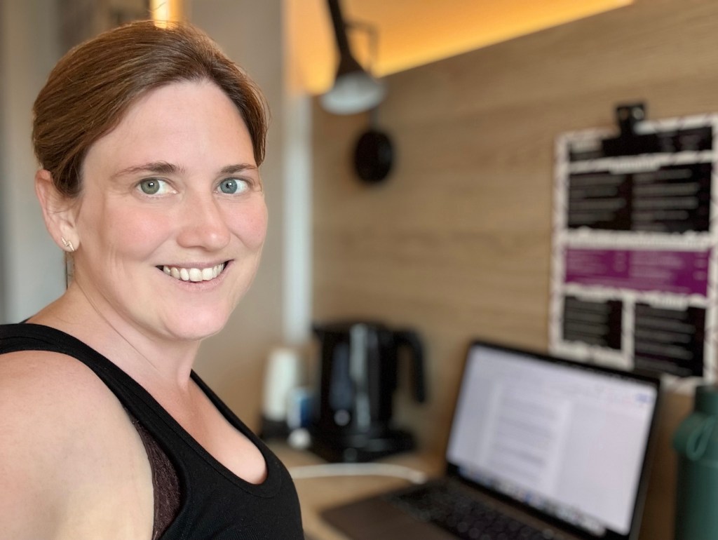 A white woman with brown hair sits at a small desk in a hotel with a laptop