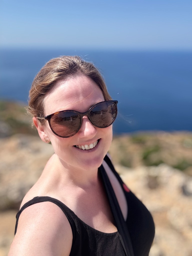 A white woman (me) with brown hair tied back. She is wearing sunglasses and is smiling.
