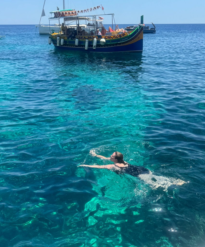 A white woman with brown hair swimming in a clear blue sea. There is a multi-coloured boat in the background