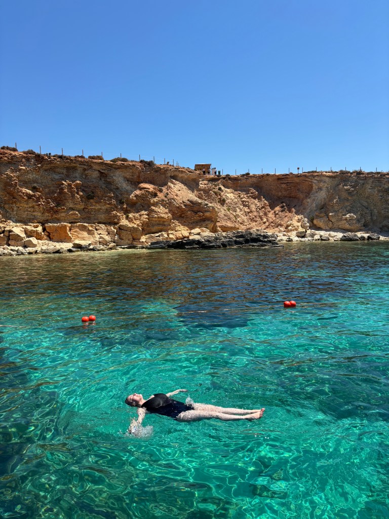 A (very) white woman floats in a clear blue sea with a rocky sandstone coast in the background and a blue sky