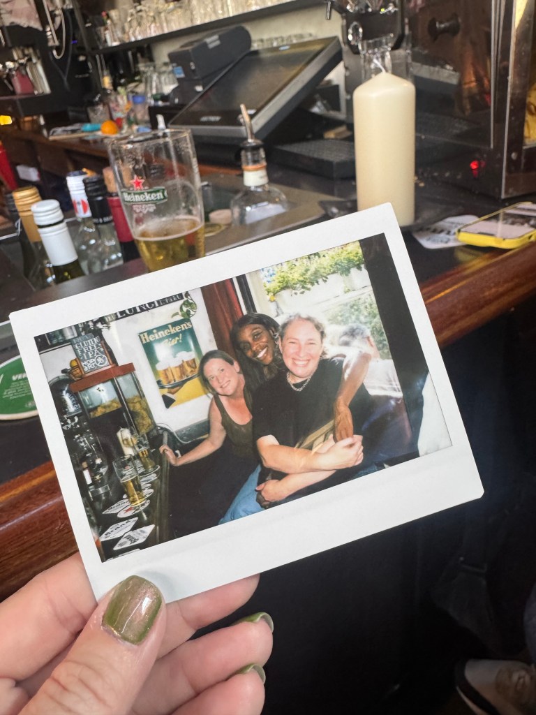 A hand holds a polaroid photo of three women in a bar.