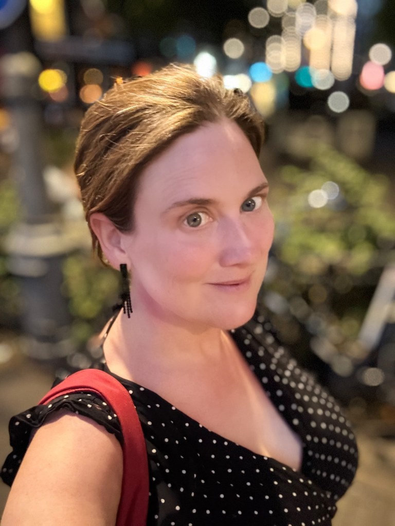 Selfie of a white woman with brown hair tied up, wearing black earrings and a black dress