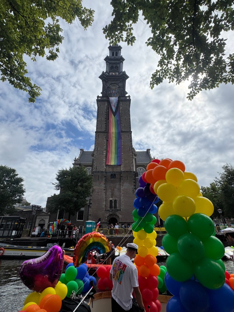 Amsterdam's Westerkerk church with a Rainbow Flag banner hung down the front. In the foreground are lots pf rainbow-coloured balloons.