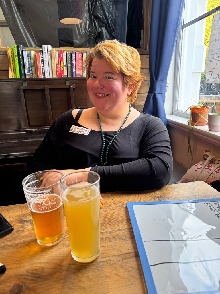 A person smiling while sitting at a table, with two glasses of beer in front and bookshelves in the background.