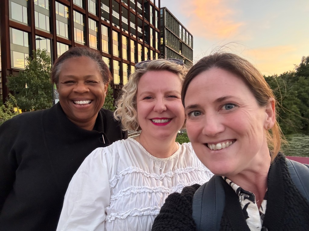 Three women smiling together for a selfie in front of a modern building during sunset.