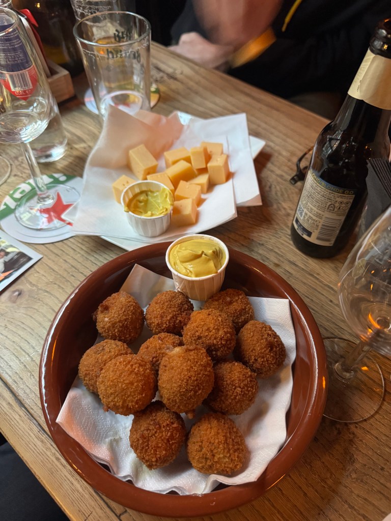 A dish featuring a portion of deep-fried croquettes, served alongside cubes of cheese and mustard, placed on a wooden table.