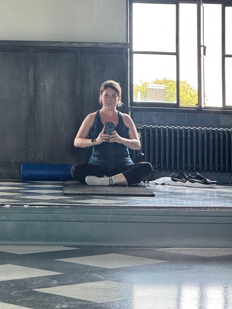 A person sitting cross-legged on a mat in a bright room, holding a phone in front of them. The background features large windows and a dark wooden wall.