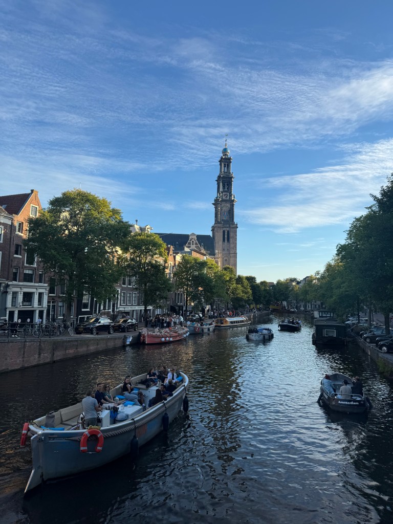A scenic view of an Amsterdam canal with boats and people enjoying the waterfront, featuring a tall tower in the background against a clear blue sky.