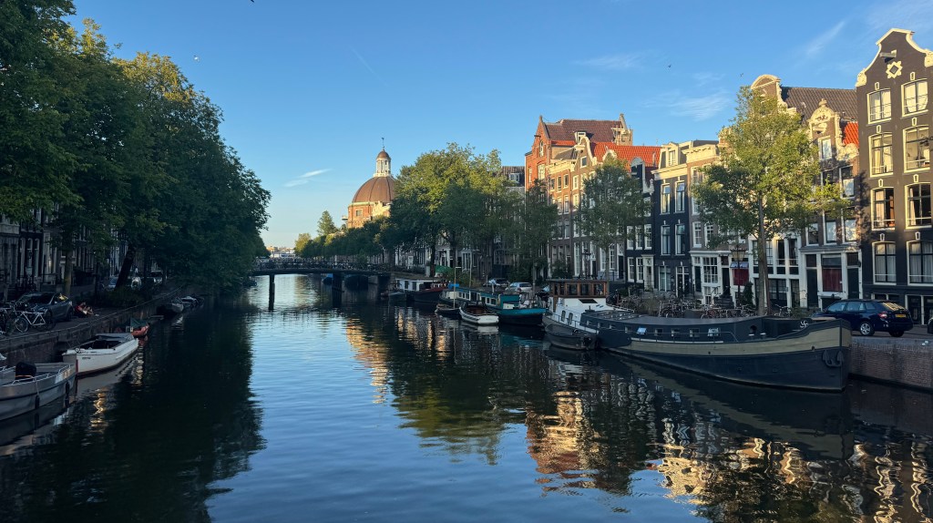 A tranquil view of a canal in Amsterdam, lined with trees and colorful buildings, reflecting in the water under a clear blue sky.