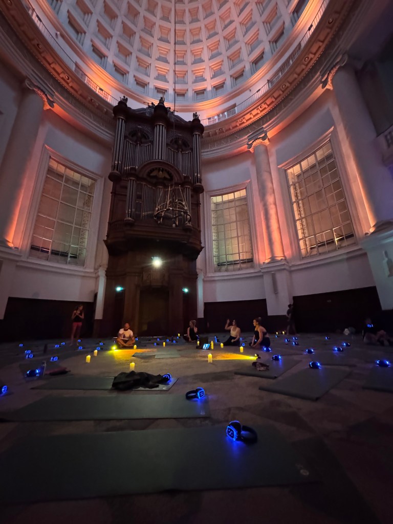A dimly lit yoga class taking place in a grand, circular room with an ornate organ. Participants are scattered on yoga mats, using wireless headphones and surrounded by glowing candles.
