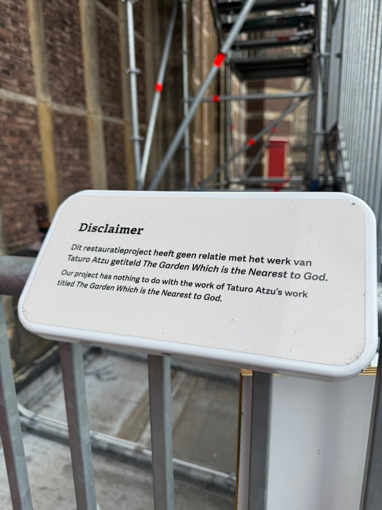 A sign stating a disclaimer about a restoration project, indicating that it has no relation to Taturo Atzu's work titled 'The Garden Which is the Nearest to God.' The sign is set against a backdrop of scaffolding and a brick wall. I have no idea why this sign exists.
