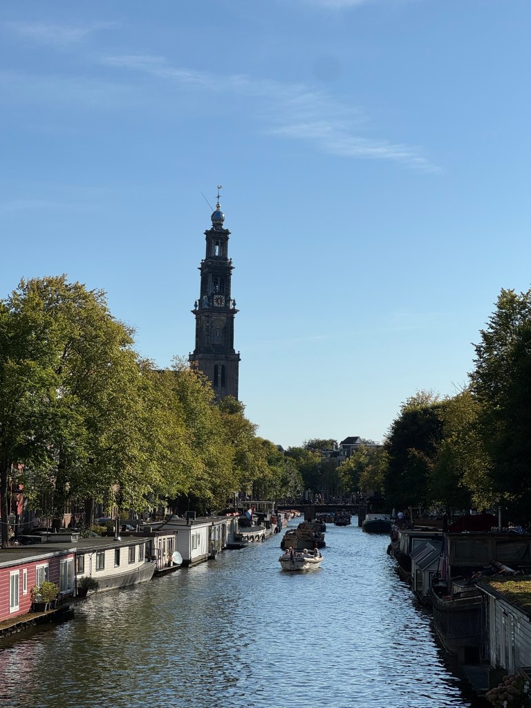 View of a canal in Amsterdam with houseboats lining the banks and a clock tower in the background under a clear blue sky.