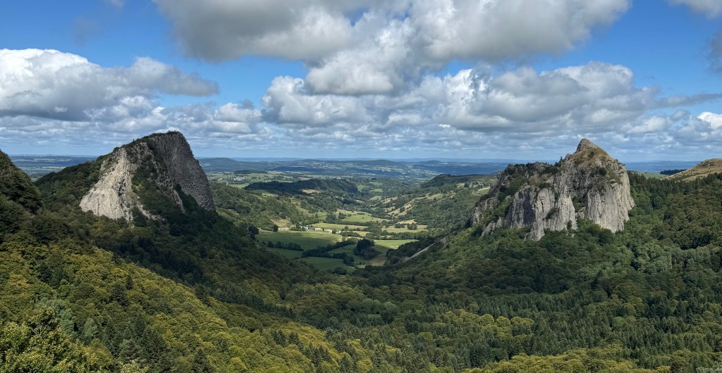 Panoramic view of a lush green valley - the Les roches Tuilière et Sanadoire - surrounded by rocky cliffs under a partly cloudy sky.