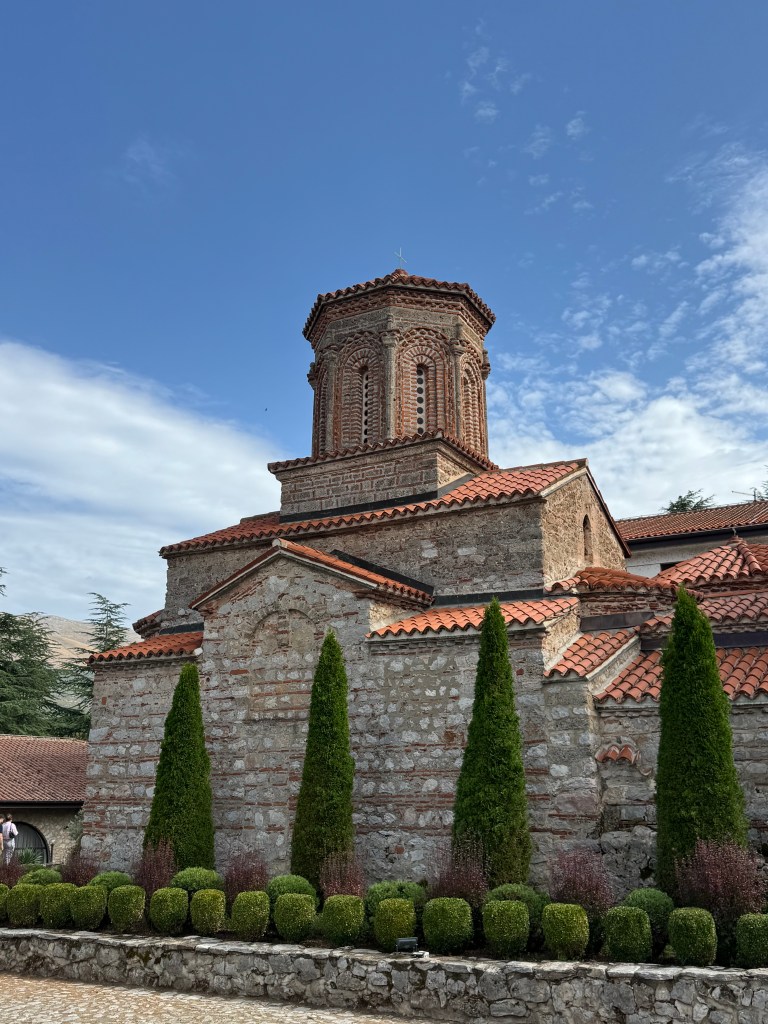 A historic stone church with a conical red tile roof, surrounded by neatly trimmed green hedges and trees, under a bright blue sky.