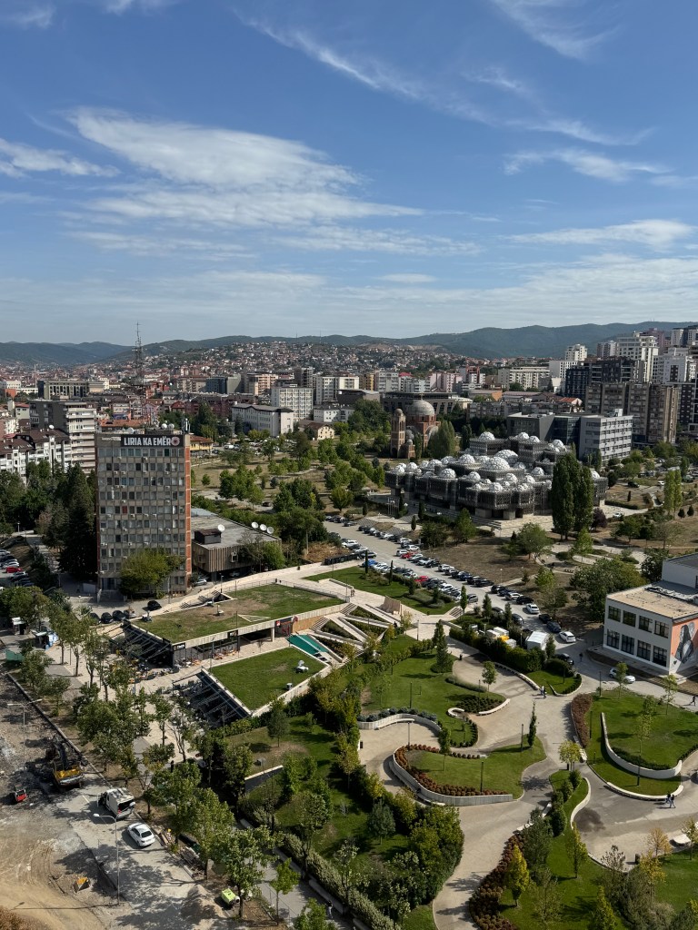 A panoramic view of a city in Kosovo, showcasing modern buildings, green park areas, and clear blue skies.