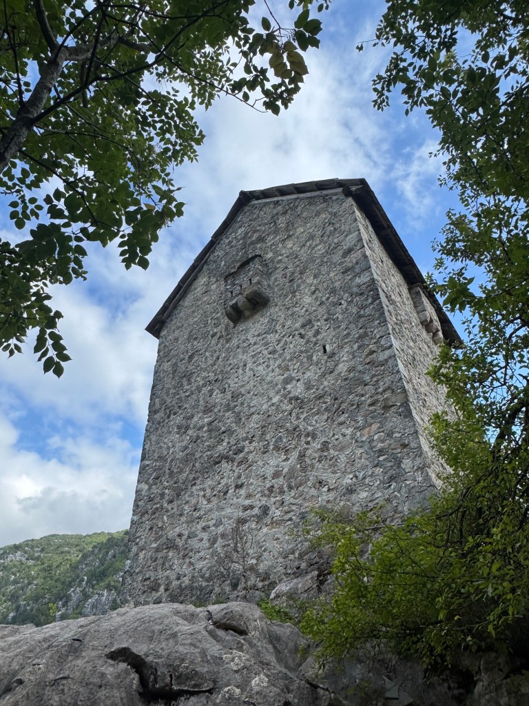 A stone structure rising from a rocky base, framed by green leaves and against a backdrop of cloudy sky.