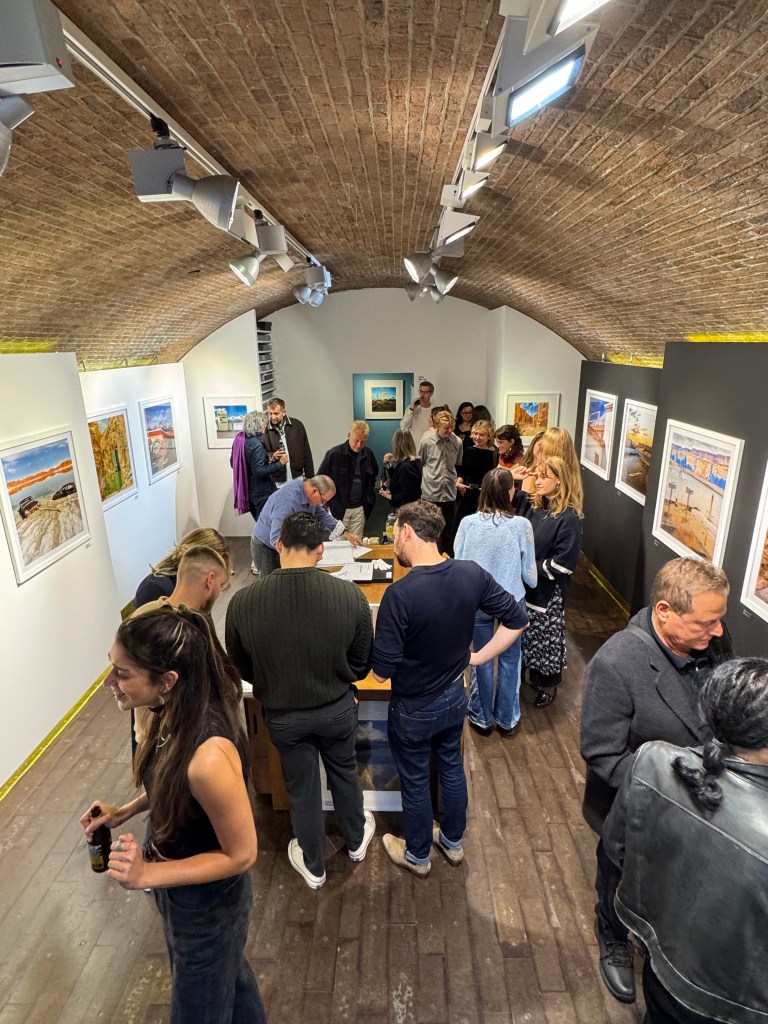 A busy art gallery exhibition featuring a diverse group of people engaging with artwork displayed on the walls, with a focus on a central table where attendees are interacting.