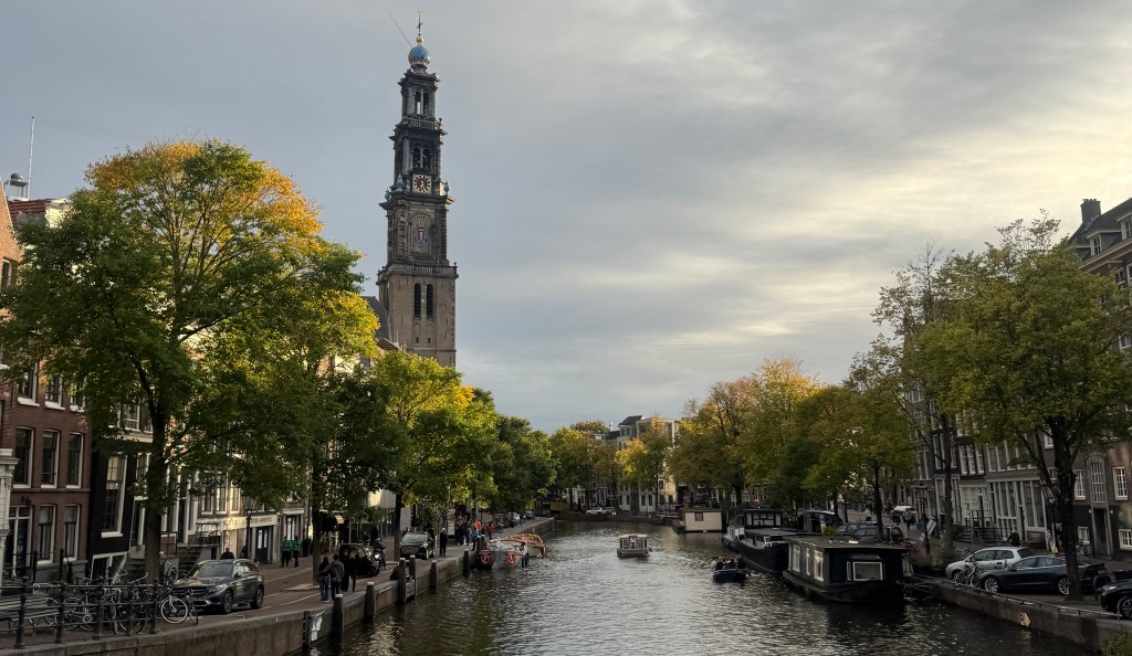 A scenic view of a canal in Amsterdam, featuring trees with autumn foliage, a clock tower in the background, and boats floating on the water.