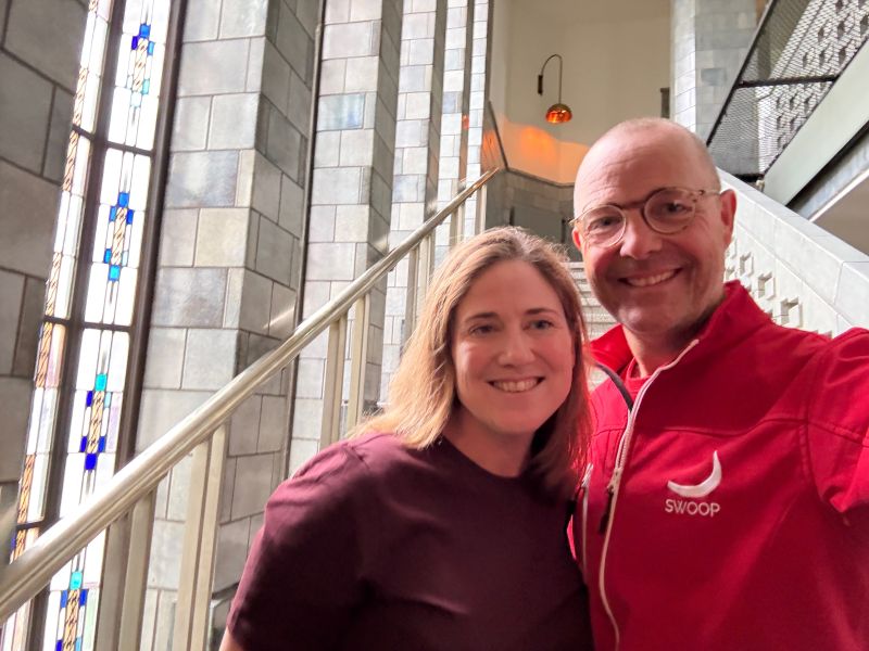 A smiling woman and man posing for a selfie on a staircase with stained glass windows in the background.