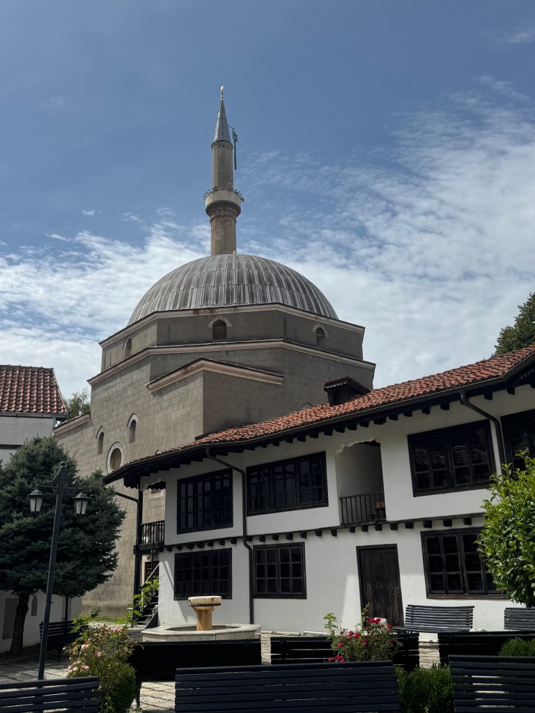 A view of a traditional architectural structure featuring a dome and minaret, surrounded by trees and a courtyard with a fountain.