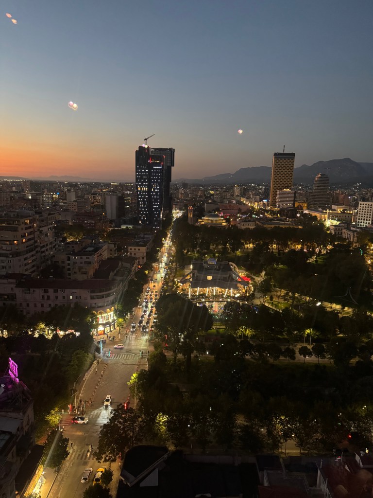A panoramic view of a city at sunset, showcasing tall buildings and lighted streets, with a mountain range in the background.