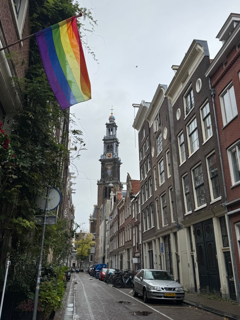 A narrow Amsterdam street lined with traditional buildings, featuring a rainbow flag hanging on the left and the tower of a church visible in the background, under a cloudy sky.