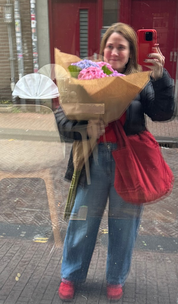 A person holding a large bouquet of flowers, wearing a black jacket and red shirt, with a red bag slung over their shoulder. They are reflected in a shop window on a street in Amsterdam.