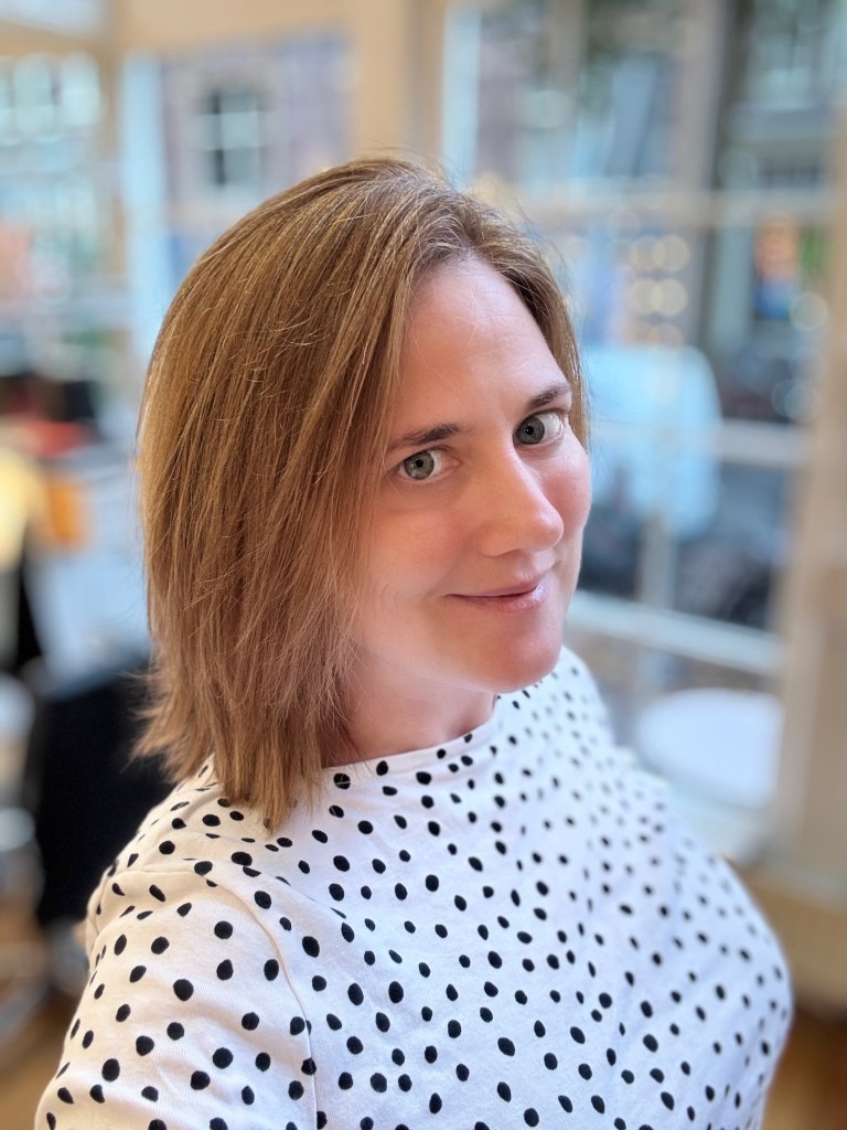 A person with shoulder-length brown hair, wearing a white shirt with black polka dots, smiles slightly while posing in a well-lit indoor setting.