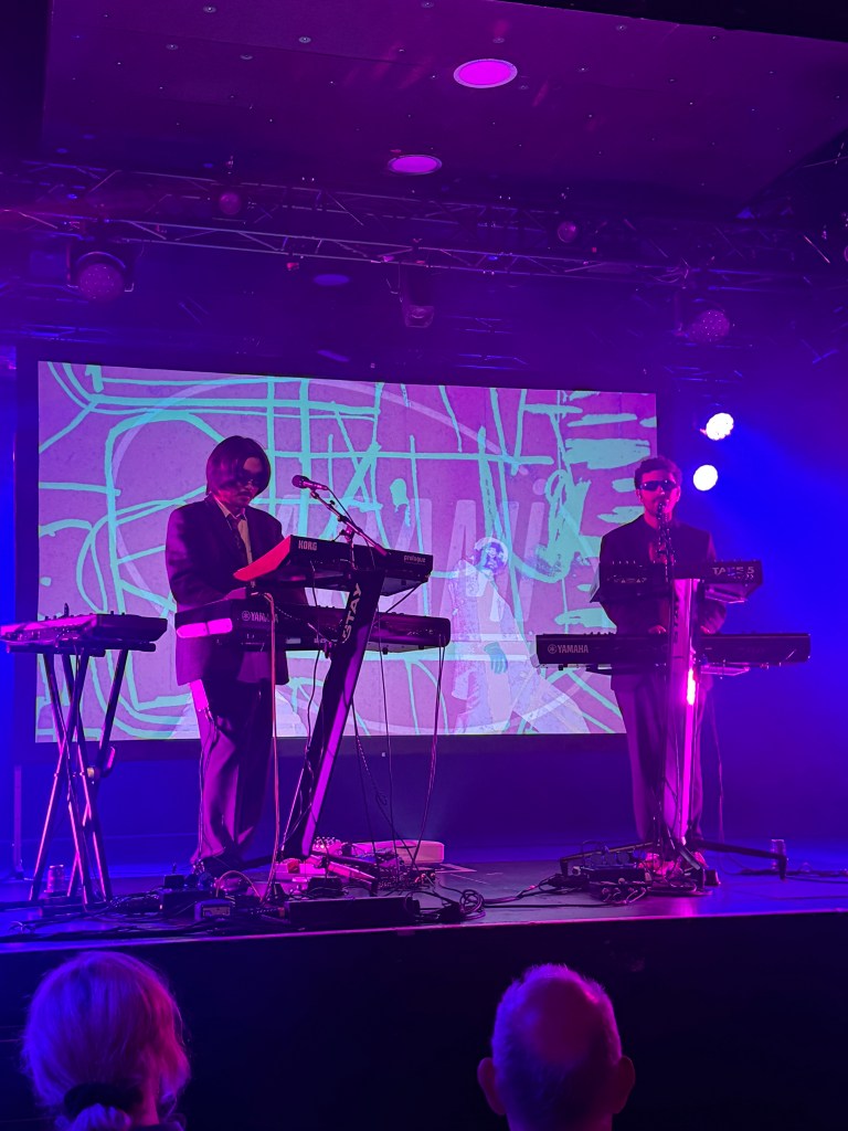 Two musicians performing on stage with keyboards, illuminated by purple lighting, and an abstract background projection.