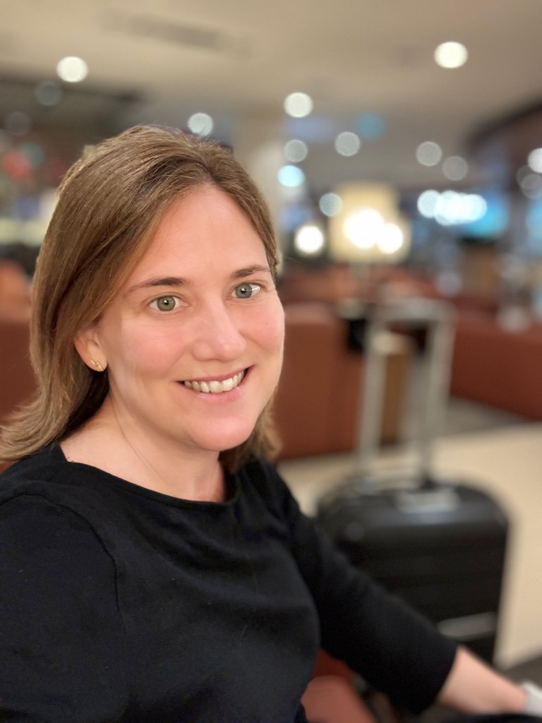 A woman smiling in a casual black top, sitting in a lounge area with a suitcase in the background and soft lighting.