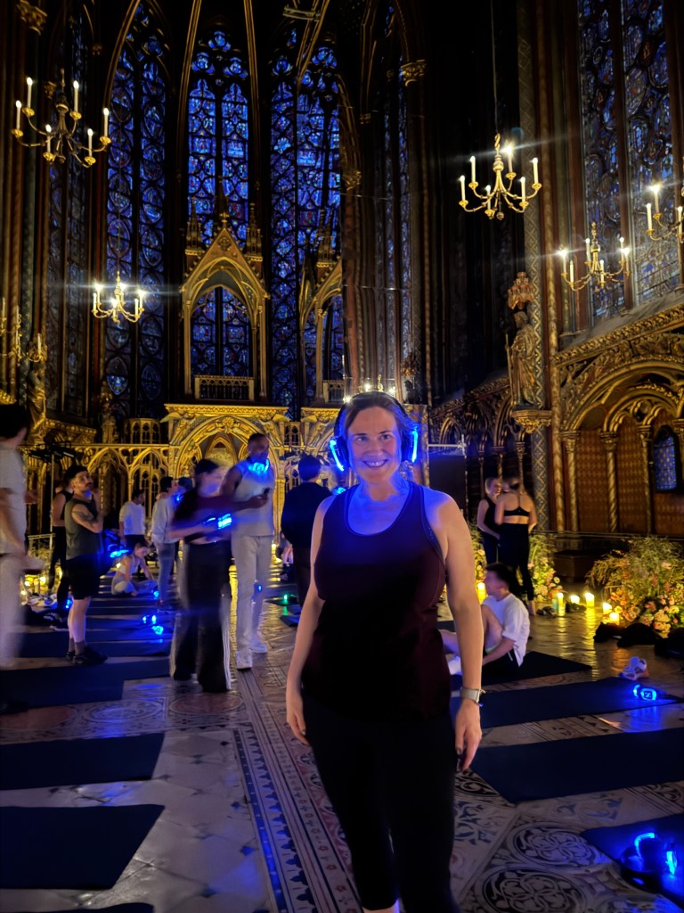 A woman smiling while participating in a dance exercise class inside a beautifully illuminated gothic chapel, with colorful stained glass windows in the background.