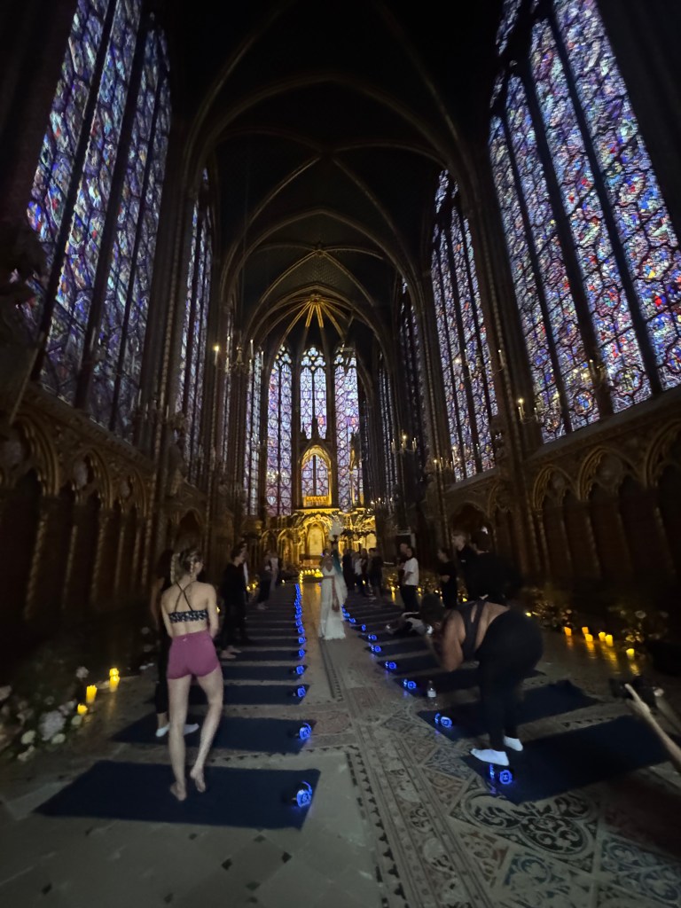 An indoor yoga class taking place in a grand, Gothic-style chapel with colorful stained glass windows. Participants are on yoga mats, and soft lighting enhances the serene atmosphere.