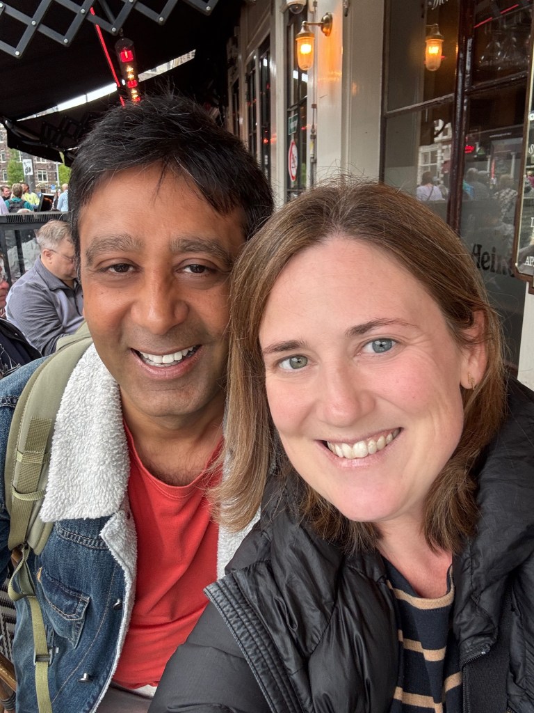 A smiling man and woman taking a selfie outdoors, with a cafe ambiance in the background.