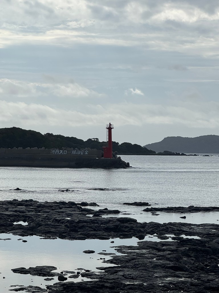 A red lighthouse standing on a rocky shoreline, overlooking calm waters with a cloudy sky in the background.
