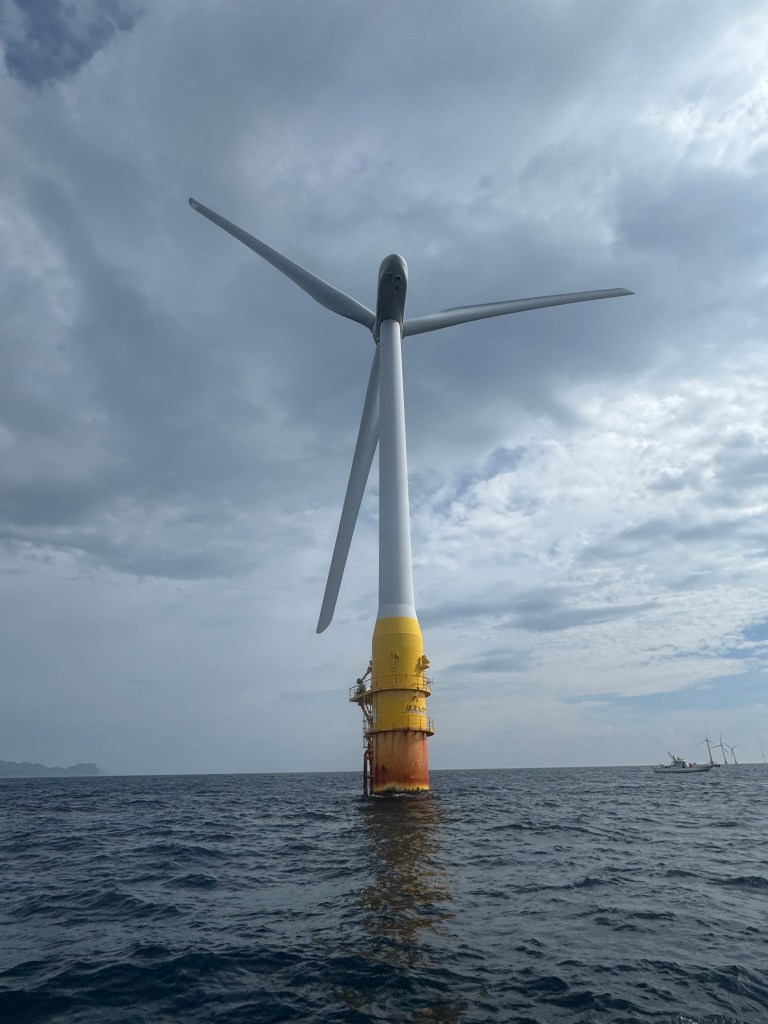 A tall offshore wind turbine with a yellow and white base, standing in the sea under a cloudy sky.