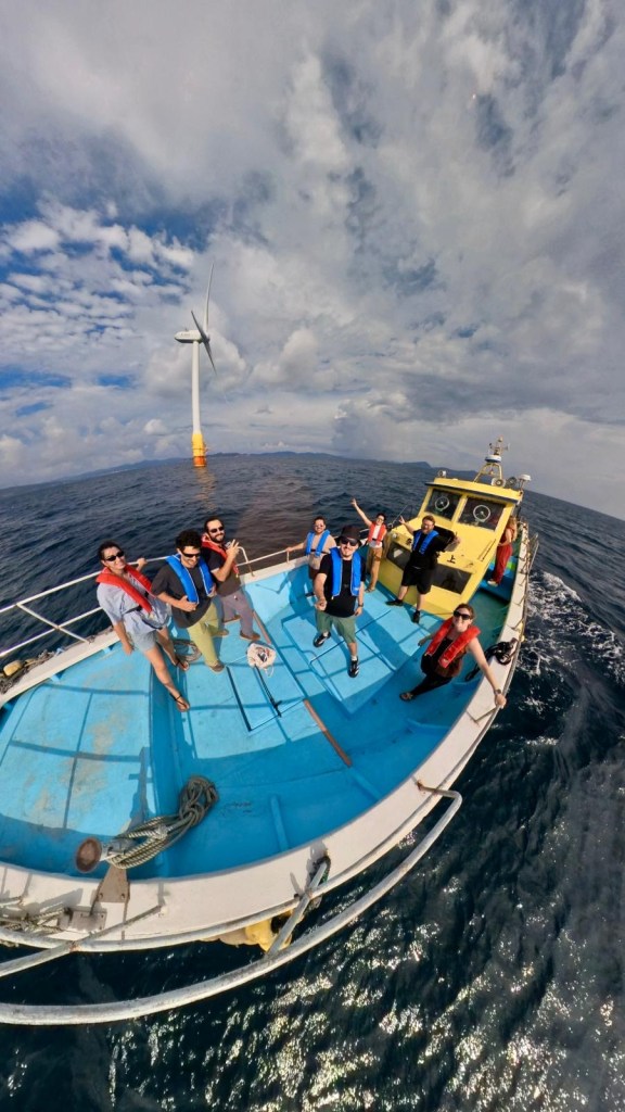 A group of people on a small boat in the ocean, wearing life vests, with a wind turbine visible in the background and a cloudy sky overhead.