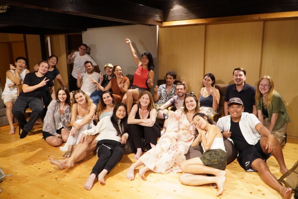 A lively group photo of diverse individuals posing together in a cozy indoor setting. Some people are smiling and making peace signs, while others are seated or leaning against each other, creating a joyful and relaxed atmosphere.
