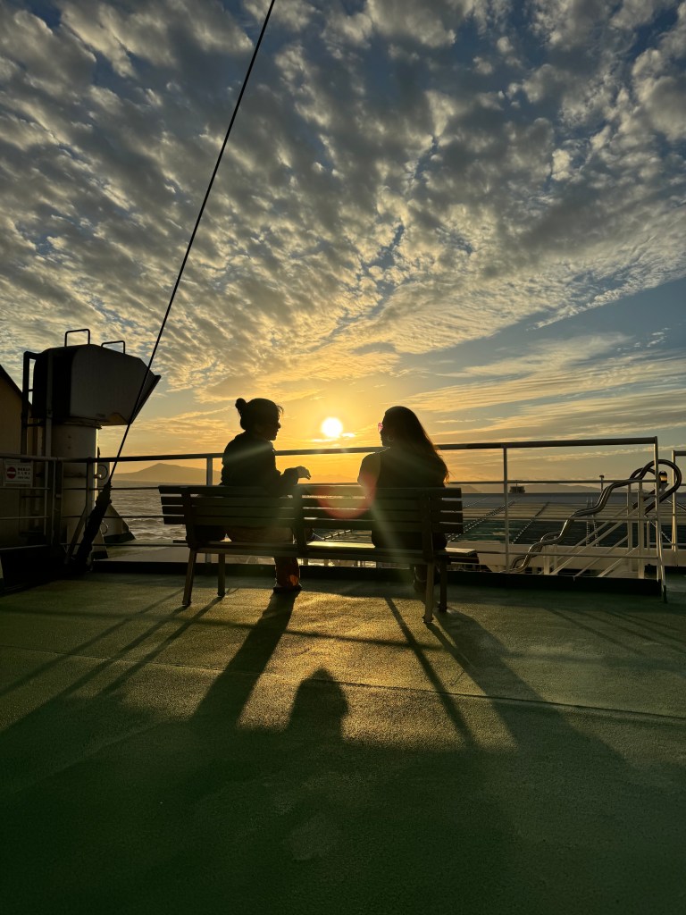 Two silhouetted figures sitting on a bench on a ferry, with a sunset and colorful clouds in the background.