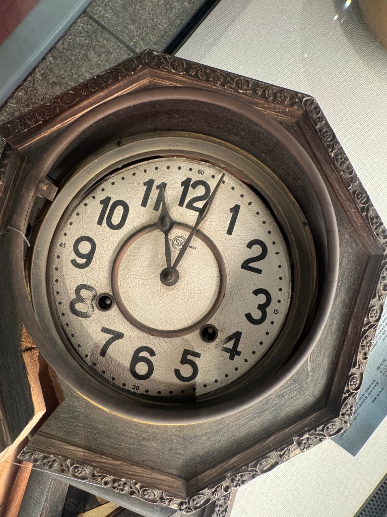 An antique octagonal clock with faded numbers and hands, displayed in a glass case. The time is frozen at 11.02 - the moment the atomic bomb hit.