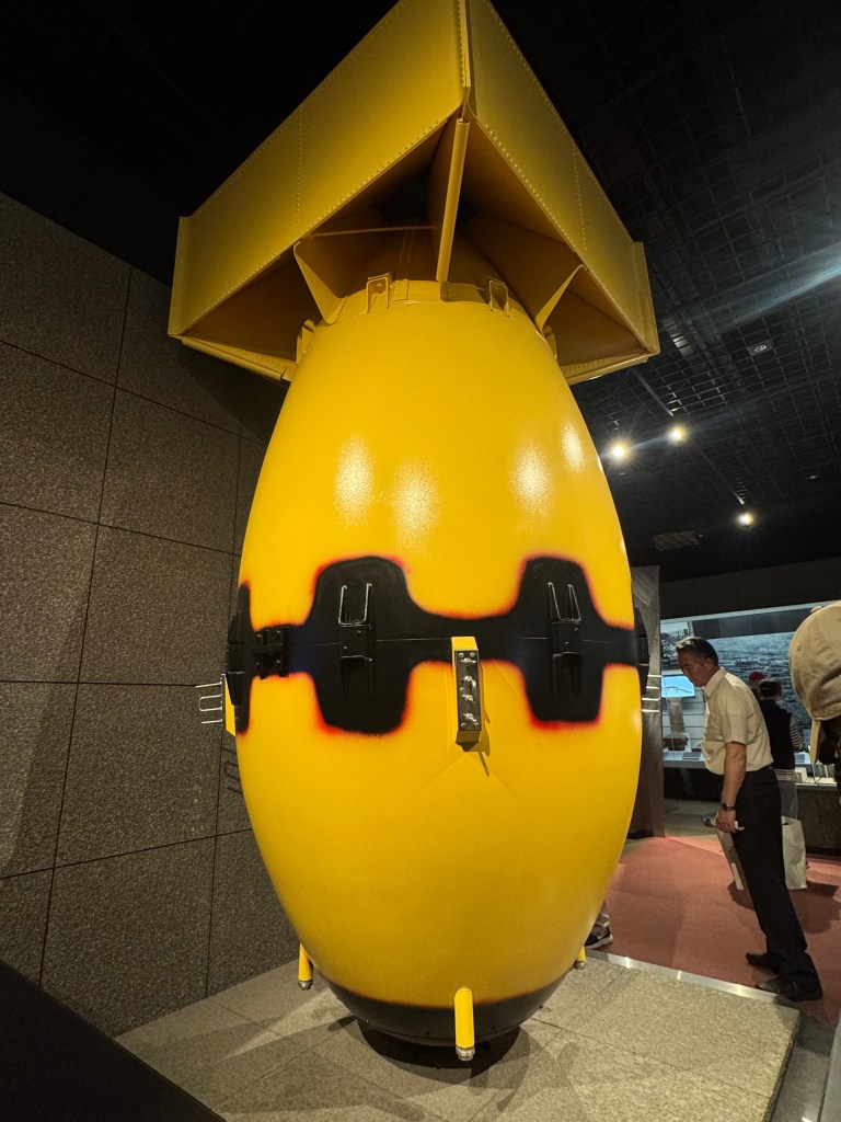 An exhibit showcasing a large yellow bomb in a museum setting, surrounded by gray walls and museum visitors.