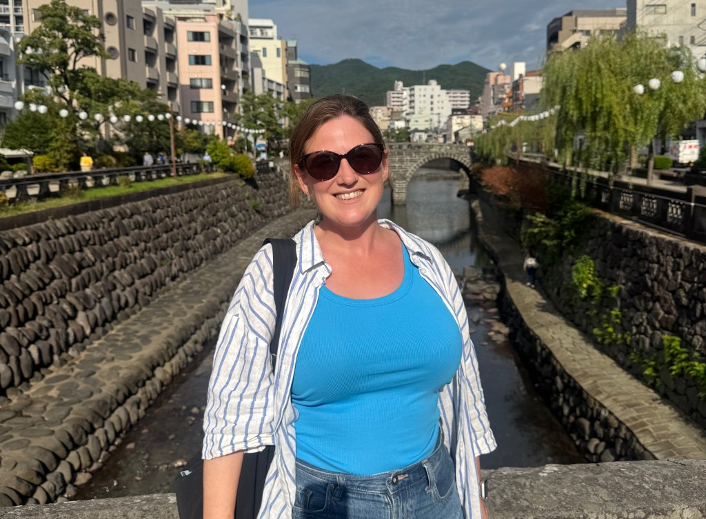 A person wearing sunglasses and a blue top stands on a bridge with stone walls, a river behind them, and buildings in the background.