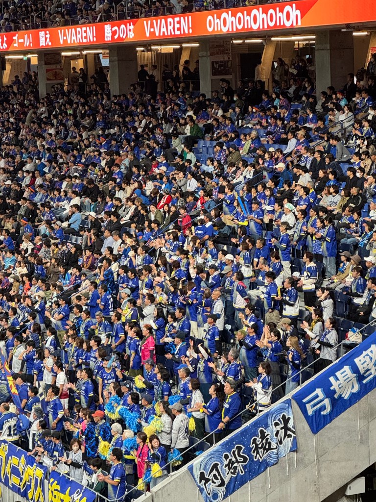 A large crowd of enthusiastic fans wearing blue jerseys cheer in a stadium, surrounded by banners promoting their team.