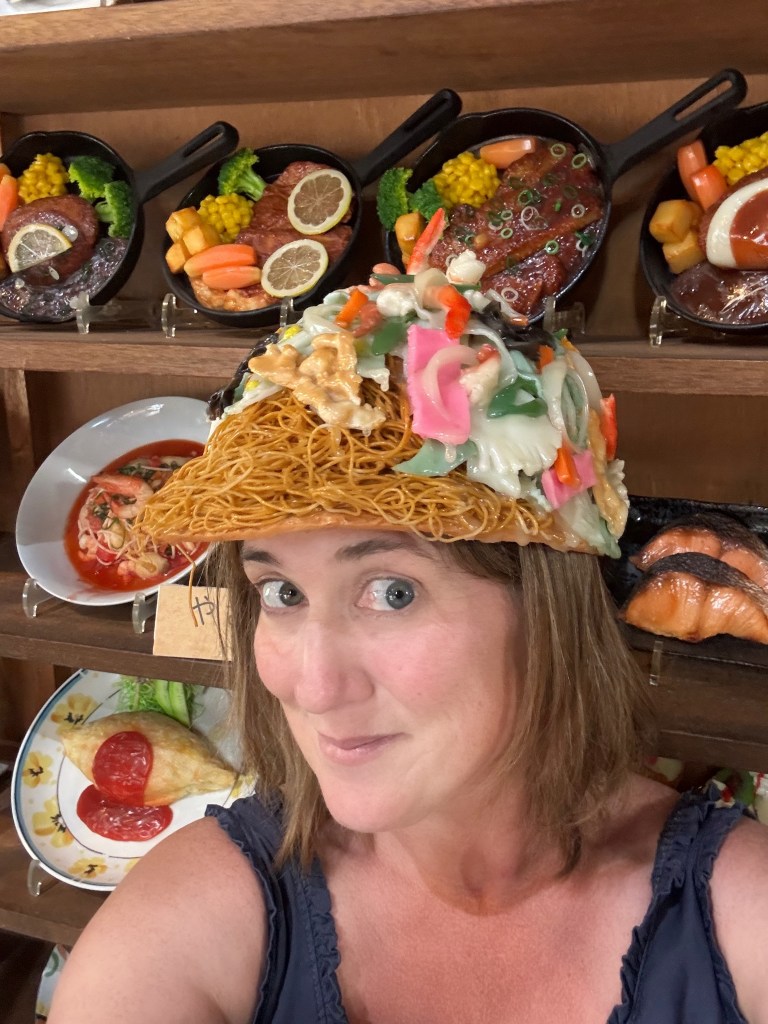 A person smiling while wearing a large replica of noodles and vegetables as a hat, surrounded by displayed fake food dishes on a wooden shelf.
