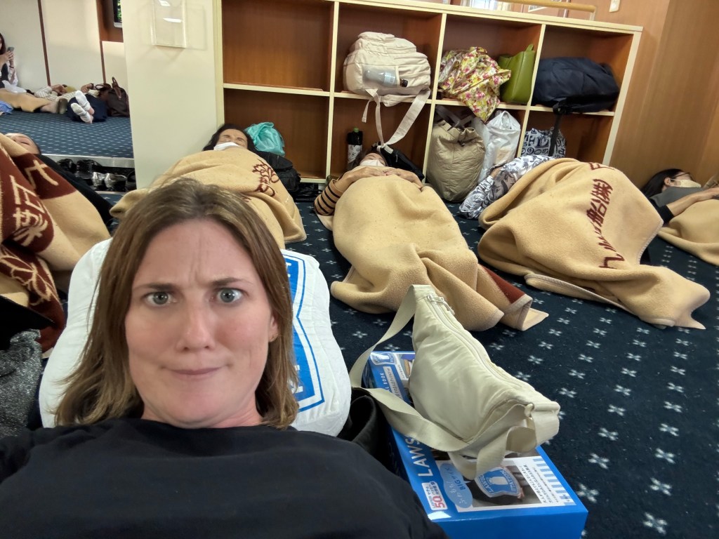 A woman lays on a pillow, looking perplexed, while others are seen resting nearby wrapped in blankets on a floor in a communal space.